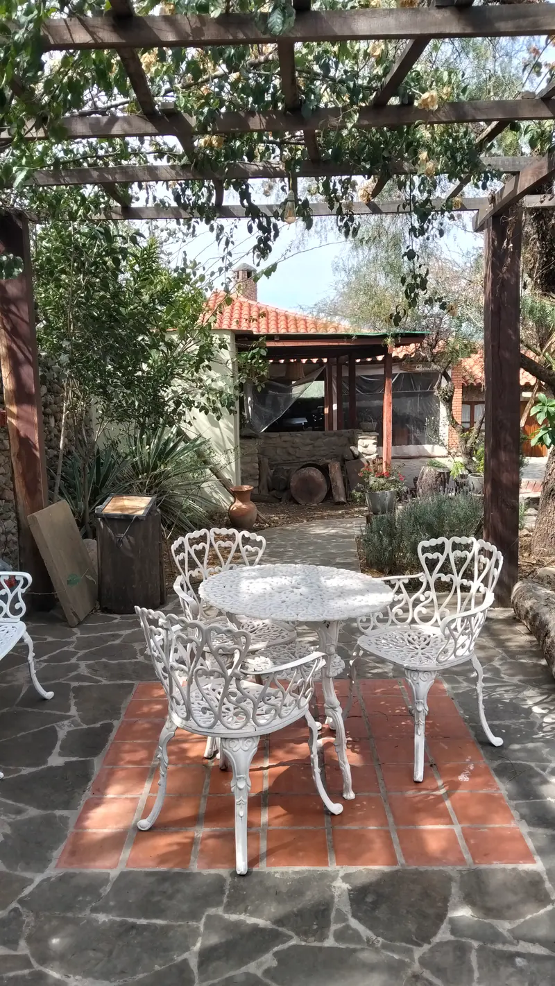 Patio con piso de piedra, mesa y sillas de hierro blanco bajo la pérgola
